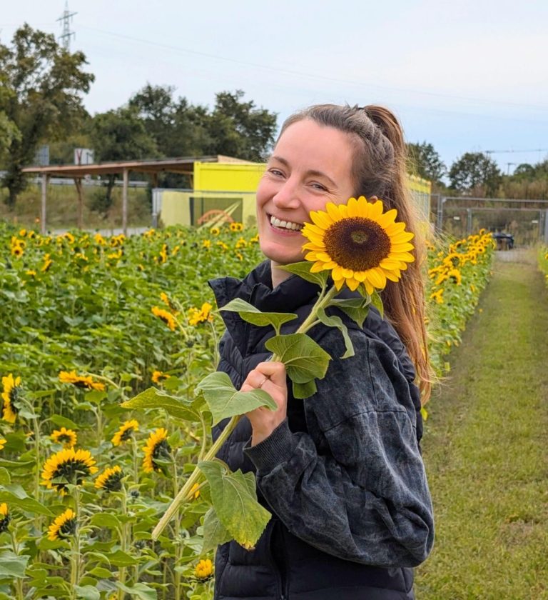 Mariana hält lächelnd eine Sonnenblume neben ihr Gesicht, während sie vor einem Sonnenblumenfeld steht.